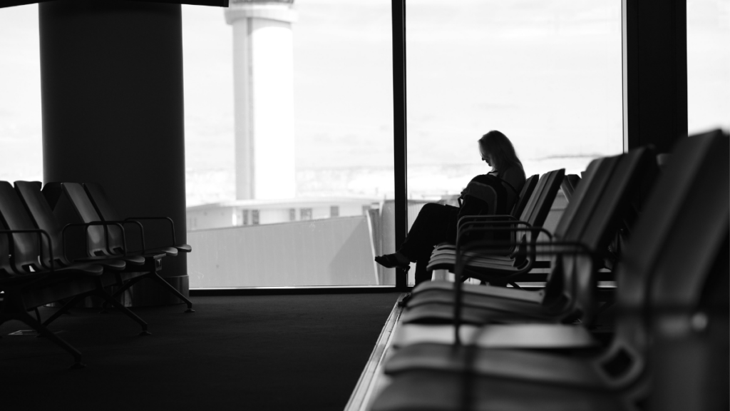 Voyageuse assise seule dans une salle d'attente d'aéroport devant une baie vitrée, illustrant le temps consacré aux trajets et aux attentes lors d'un voyage.