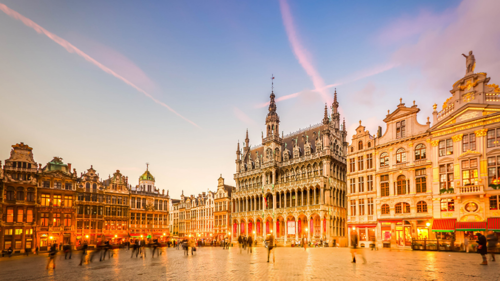 Grand-Place de Bruxelles au coucher du soleil, avec l'Hôtel de Ville illuminé et plusieurs passants sur la place pavée.